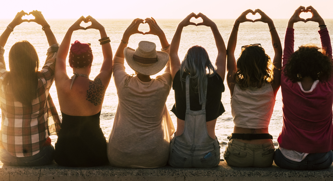 Six women sitting together looking at the ocean with their hands in a heart shape